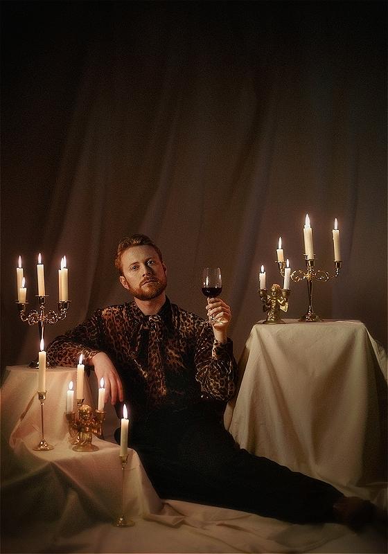 John is lying luxuriously on a draped surface, wearing leopard-print shirt and tie. They are holding up a glass of red wine and are lit by elegant white candles on multiple candlesticks and candelabras around them.