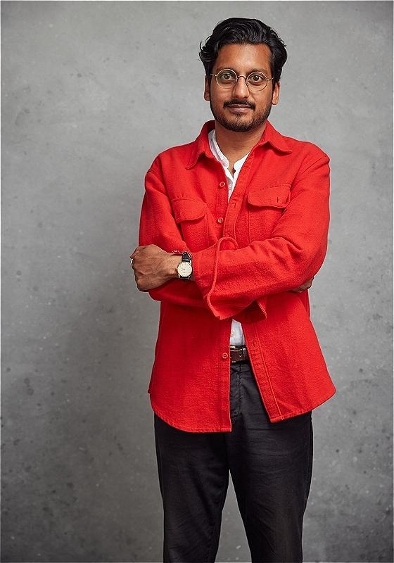 Ahir stands with their arms crossed over their chest, looking at the camera. They're wearing a bright, salmon-coloured top.