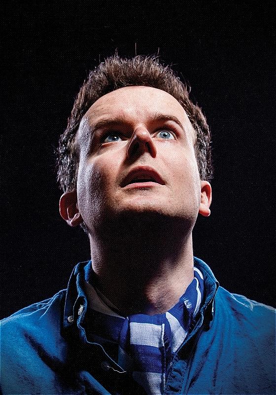 Andrew is looking up and behind the camera with a somewhat shocked look on their face. They're wearing blue against a pitch black background.