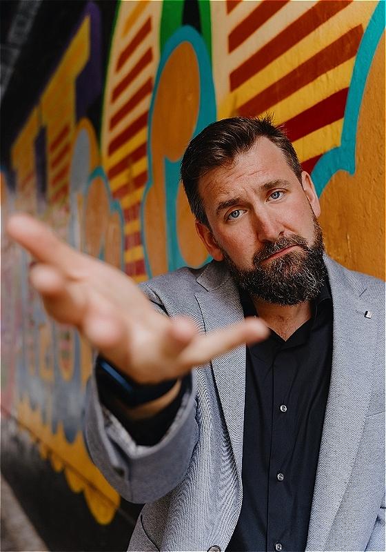 Garrett, wearing a grey suit, offers the camera his hand, before a wall with colorful paint.