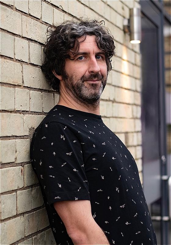 A person in a black top leans back against a brick wall and looks into the camera. They have dark curly hair, a dark beard and light skin, and they are wearing a black T-shirt. There appears to be a tree in the foreground just off to the side. 