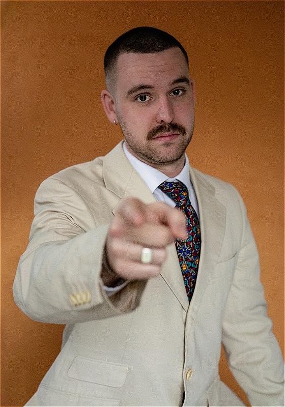 Rob is wearing a cream suit and a colourful tie against a russet background. They are pointing directly at the camera.