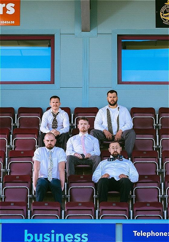 Five men wearing white button down shirts and ties sit on red bleacher seats staring straight ahead seriously.