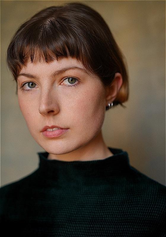 Headshot of the performer wearing a dark turtleneck sweater before a beige background.