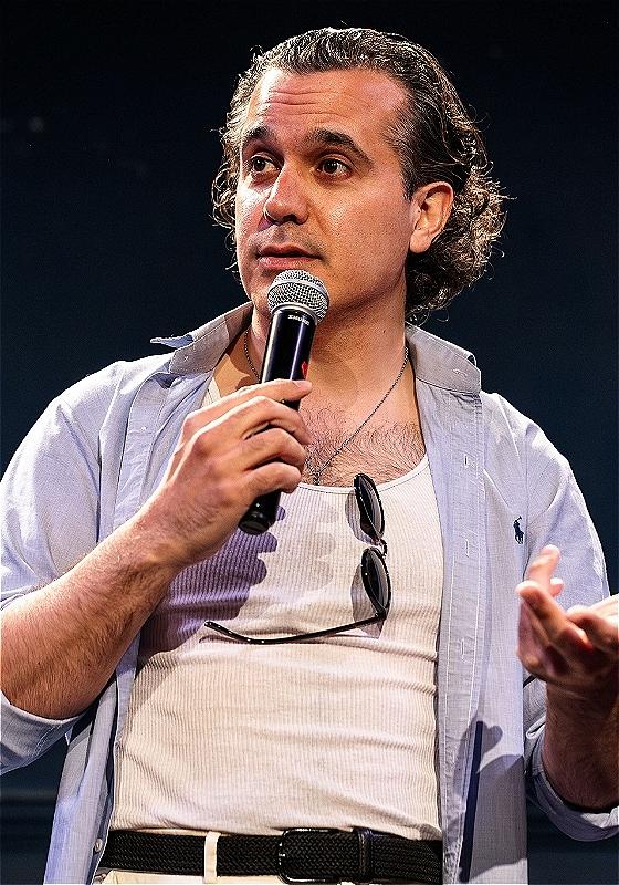 Sami hold a mic up to their face while delivering a set.  They've got sunglasses hooked over a tank top underneath an open shirt. 