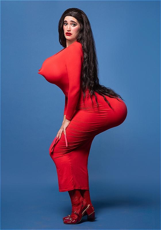 Maya is posing sensually in a red dress, with comically and alarmingly oversized padding on their posterior and chest. They're rocking a strong red lip and a black wig, which spills over the shelf formed by their amplified posterior.