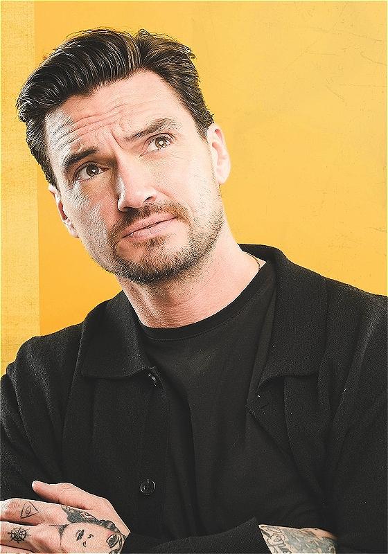 Jack is crossing their arms and looking off-camera. They are wearing black which contrasts with the bright yellow background. Their hands are tattooed - one of these tattoos appears to be a cartoon face.