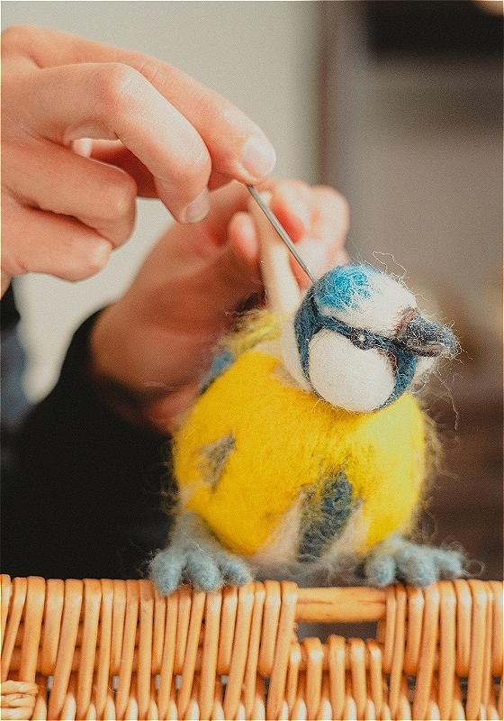 A pair of hands manipulate a blue and yellow bird made of string, on top of a wooden perch.