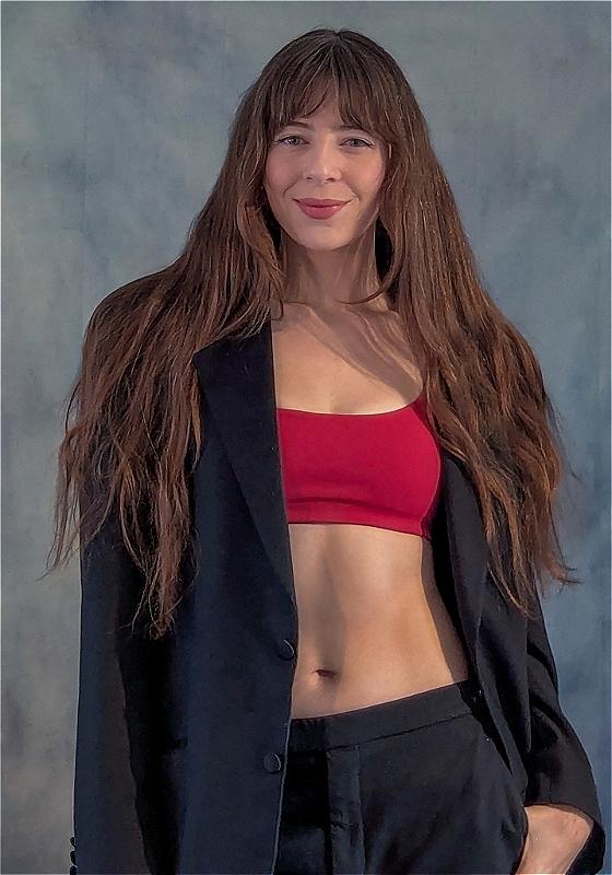 Chelsea, wearing a red sports bra over a black blazer, smiles at the camera before a grey background.