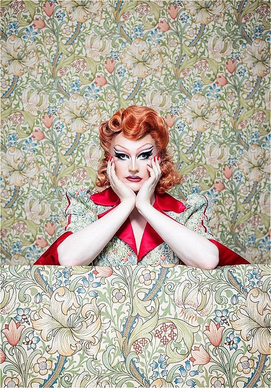 Drag queen Kate Butch stares ahead while wearing a flowery pattern dress that merges into the background, which has the same pattern.