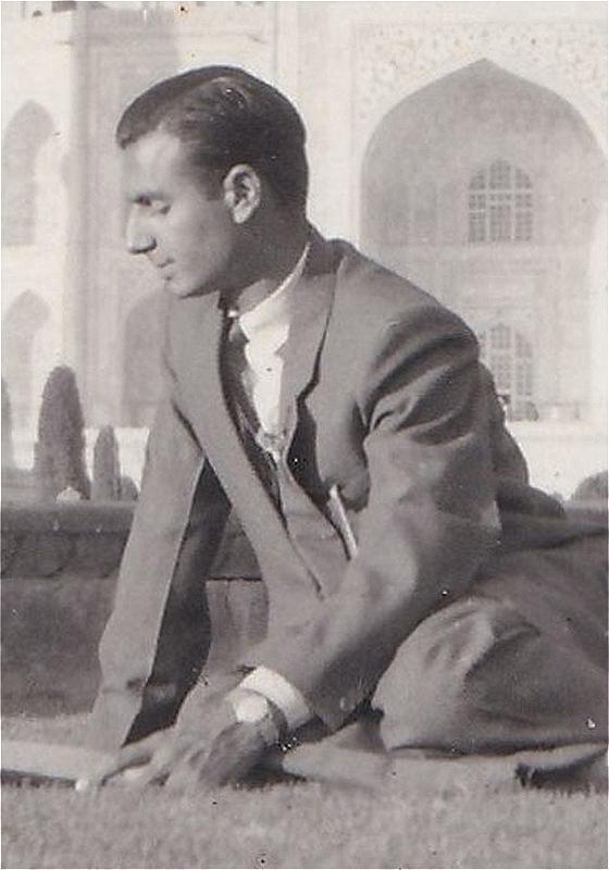 An old black and white photo of a person in a suit. Their eyes are closed and they appear to be sitting on some grass, with what looks like a fortress in the background. It has beautiful arches.