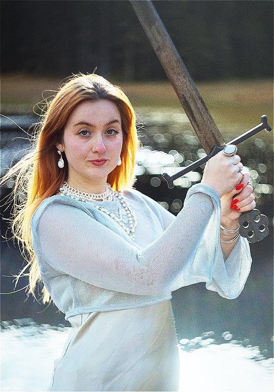 Em is wearing diaphanous white clothing and weilding a broadsword. There is a body of water in the background. They look ready to throw down.