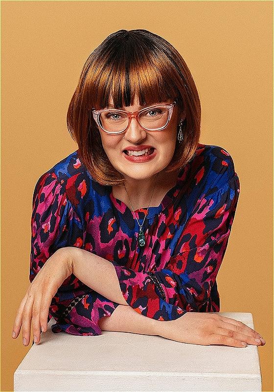 Emmeline is leaning towards us conspiratorially with their arms crossed along a white surface. They're wearing a blue and pink leopard print top and have got a fabulous bob and sparkly glasses. 