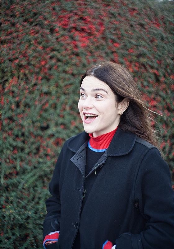 Davina is outside in a coat with their hands in their pockets, They're smiling at something off camera