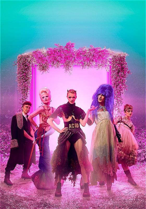 Five artists in fabulous costumes of all colours, including one especially bodacious purple wig, stand in front of a glowing pink archway portal, which is wreathed in flowers, possibly lilacs. They are all striking dynamic poses. 