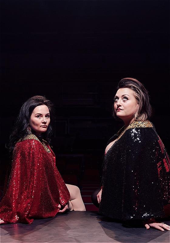 Two people sitting on the edge of a stage, in a darkened auditorium. We are looking at them from behind - they're twisting back round to look at the camera. They are both wearing sparkly glittery gowns in black red and gold