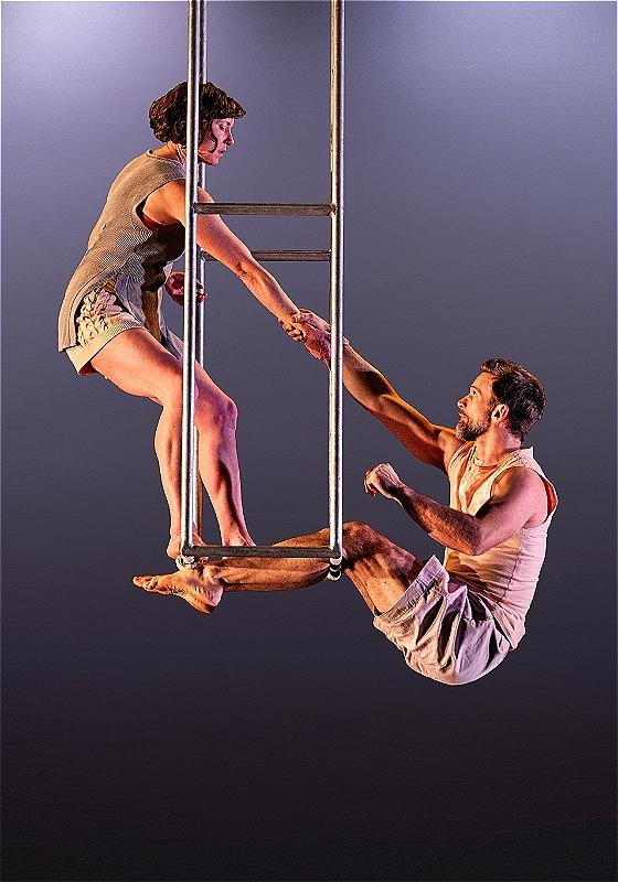 Two performers hold on to each other on an iron platform that hangs from the ceiling before a grey purple background.