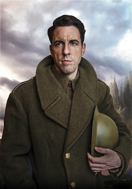 David William Bryan stands with a determined look on his face, wearing a world war one military uniform overcoat, with a helmet under his arm. The sky behind him is cloudy and moody.