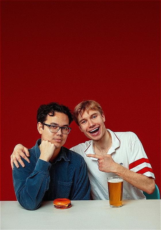 Two people sit companiably at a white surface, against a white background. One has an arm around the other, and is laughing and pointing in a friendly way as they both look to the camera. In front of them, you guessed it: one burger, and one pint. Both these items look a little disappointing in a comedic way. 
