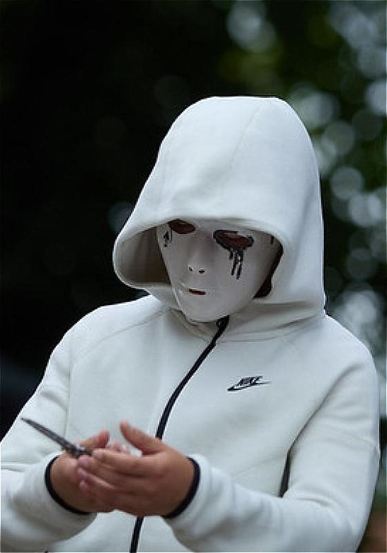 A person in a white Nike tracksuit stands with their hood up. Their face is completely covered by a mask which has black tears painted underneath the eyes. They appear to be holding something - possibly a knife