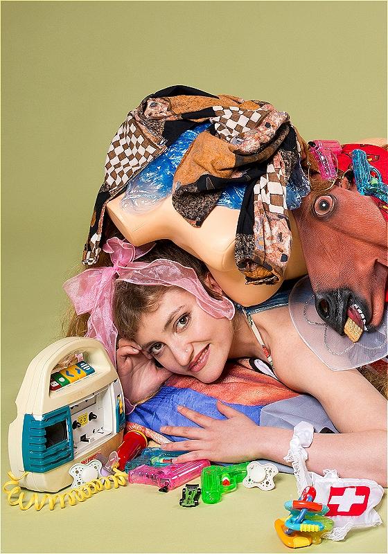 Frankie lies under a heap of random plastic tat, which includes a horsehead mask and bits of a mannequin. Beside them is an assortment of water pistols and a cassette player with a tiny plastic mic attached to it. They are smiling cheekily at the camera. 