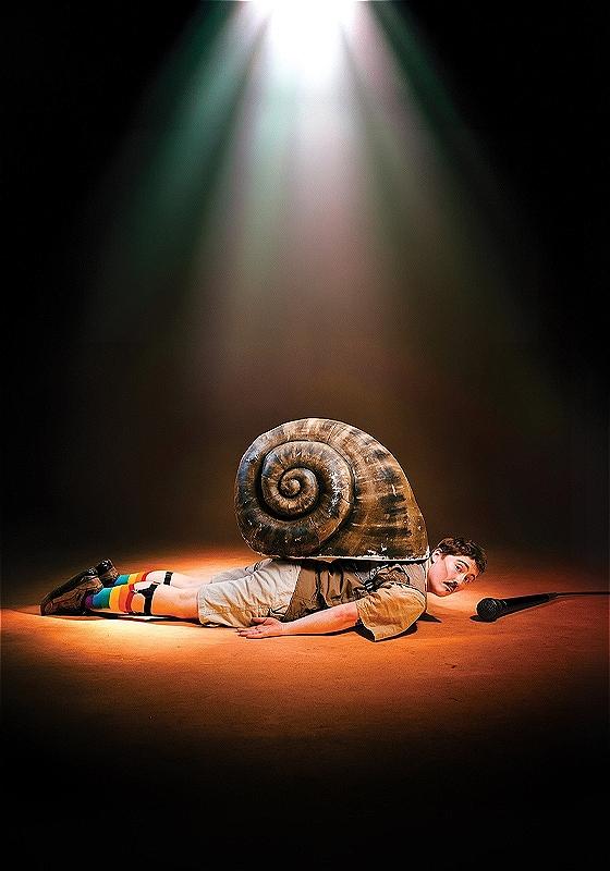 George lies on the floor under a spotlight, on their belly, with a huge snail shell on their back. they're wearing fabulous rainbow socks held up by old-fashioned sock garters to complete an otherwise beige outfit. They're looking incredulously at the camera, and a mic is on the floor beside where their head is.