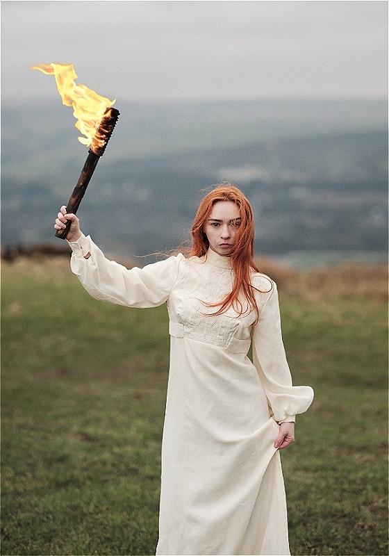 Hannah is wearing a long flowy white dress in a field, and is holding up a burning torch. They have a look of determination on their face, and their long bright red hair is dramatically blown about by the wind