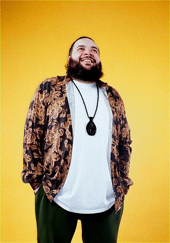 Photo of beaming Raj Poojara wearing a white shirt, a patterned overshirt, green pants, and a black necklace before a yellow background.