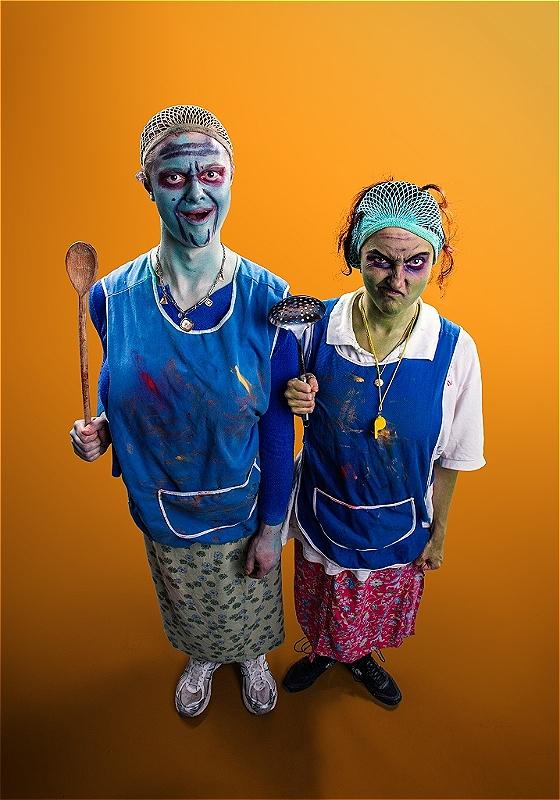 Two performers wearing blue cafeteria aprons, hair nets, and unconventional green and blue makeup hold up kitchen utensils before an orange background.