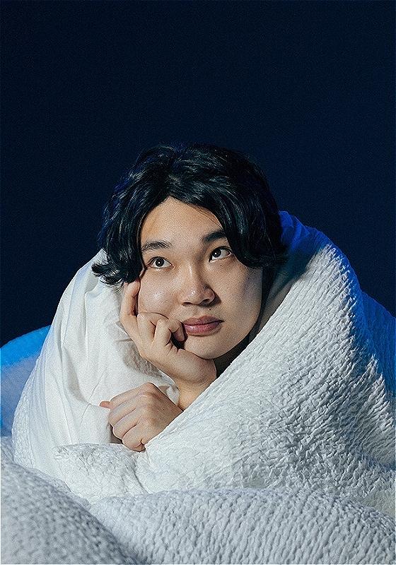 Li Jin Hao is snugly wrapped in a thick duvet, looking ponderously past the camera with their chin resting on their hand, which is all that extends from the duvet..