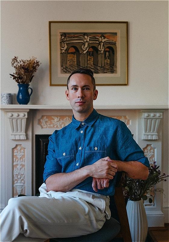 A man wearing a blue button-down shirt and white pants sits before a white living room with a fireplace, a painting and a blue vase over it.