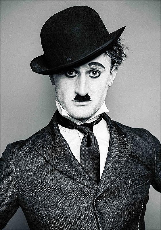 A black and white image of the performer dressed as Charlie Chaplin with a bowl hat, jacket, and tie, before a grey background.