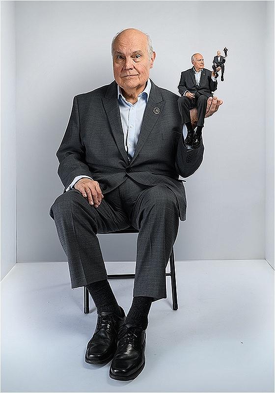 Mike sits in a chair in a small white box of a room. He's holding up a hand where a miniature of them sits, who in turn is holding up a hand that's holding another still smaller miniature twin, and so one for at least two more iterations. 