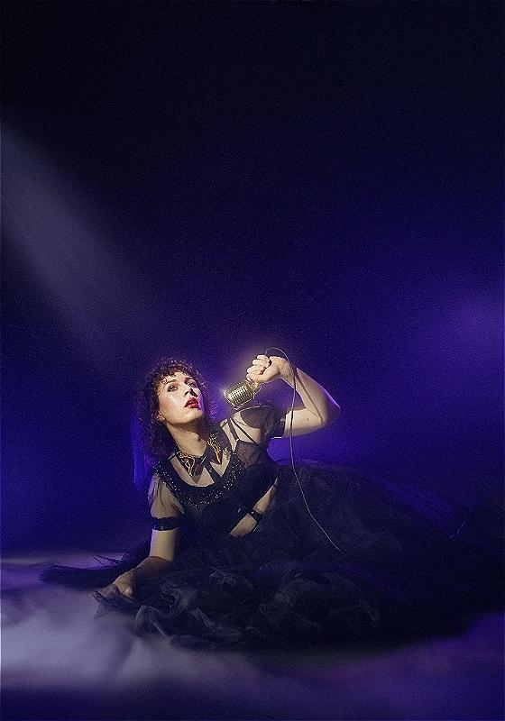 Sam in a sheer black dress lounging on the floor holding a vintage microphone. There is a purple smoky filter over the image.