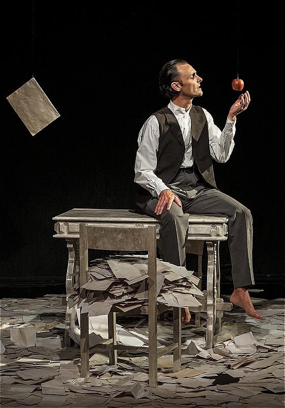 A man throws an apple as he sits on a table surrounded by countless pieces of paper, before a black background.