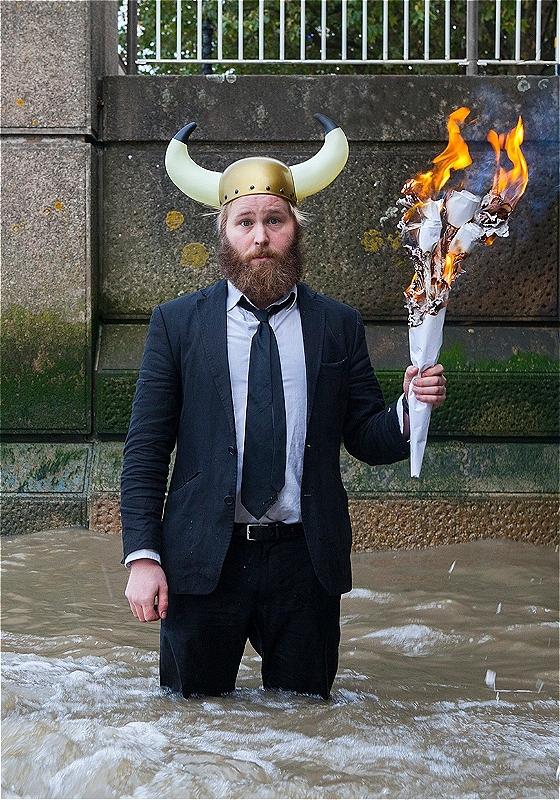James stands knee-deep in a river, wearing a suit and a cartoony viking helmet. He holds a flaming torch while looking sadly at the camera.