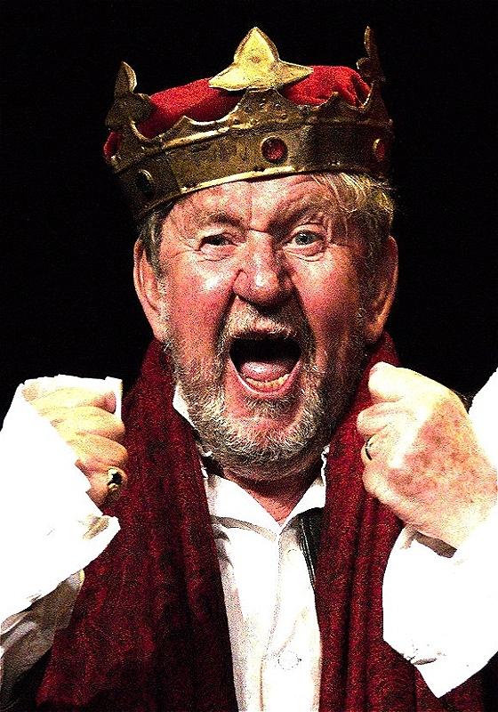 Photo of passionate Pip performing with a crown on his head, both hands raised as fists, before a black background.