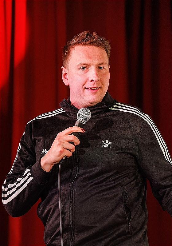 Joe Lycett wearing a black Adidas jacket and holding a microphone in front of a red curtain.