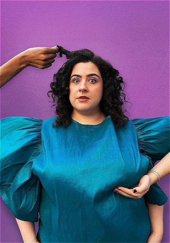 Jess in a big blue chiffon top with one hand from out of frame lifting her hair and another hand cupping her breast.