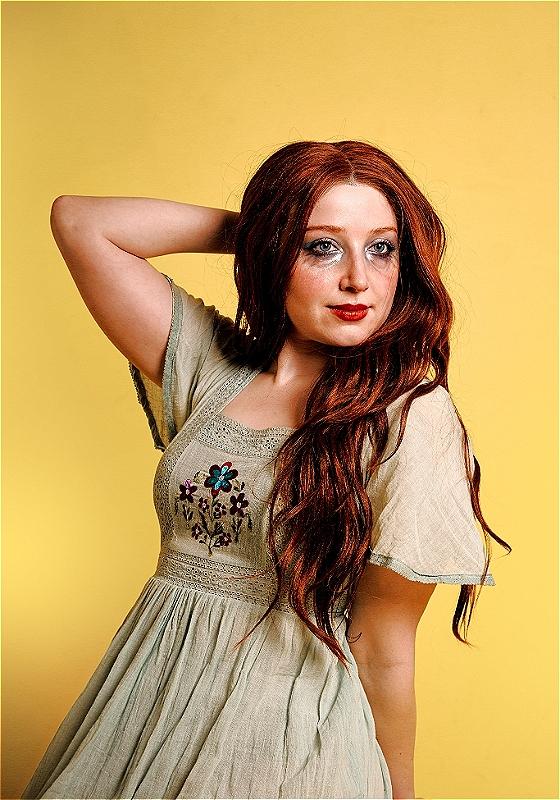 A woman with smudged makeup and wearing an old nightgown poses in front of a yellow background.