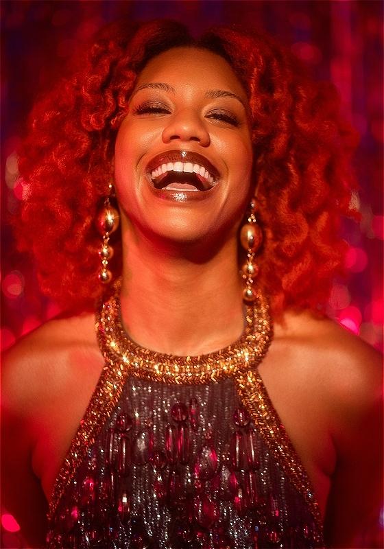 Kemah is wearing a gold sparkly halter top with matching earrings with a big laughing smile on their face. Their hair is red and matches the background.