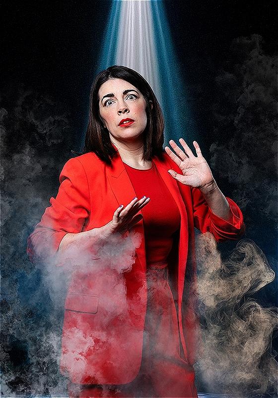 Rosie stands under a spotlight with a wide-eyed look at the camera. They are wearing a orange-red suit and are cloaked in fog.