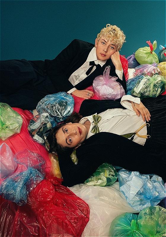 Two people in suits and bowties lies across a heap of rubbish in different-coloured refuse sacks. They're looking directly at the camera