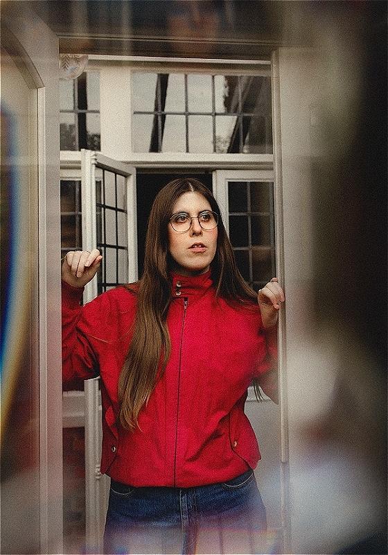 Ele, wearing a red jacket and jeans, stares out of a house, windows behind her.