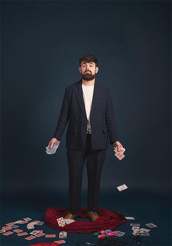 Tom Brace wearing a suit and holding a pack of cards in either hand. On the floor, several more cards and various props.