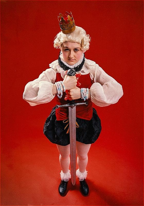 Lil standing holding a comically large sword and a small crown perched on their head like a petulant child.