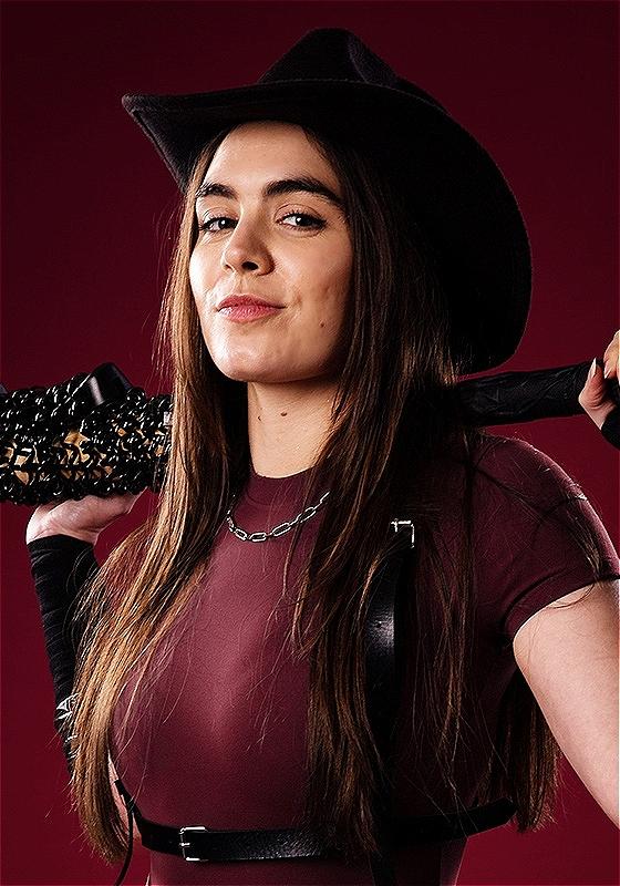 Amelia holds a baseball bat covered in barbed wire wearing a black leather harness and cowboy hat.