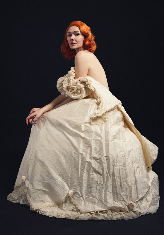 Elf sitting demurely in a unzipped floor length ballgown against a black backdrop.