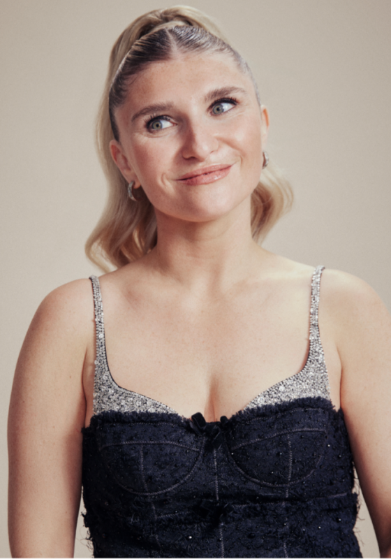 Sophie Garrad is standing against a pink background wearing a sparkly top. She's got her hair up in a slick ponytail and is smiling, looking off to the left.