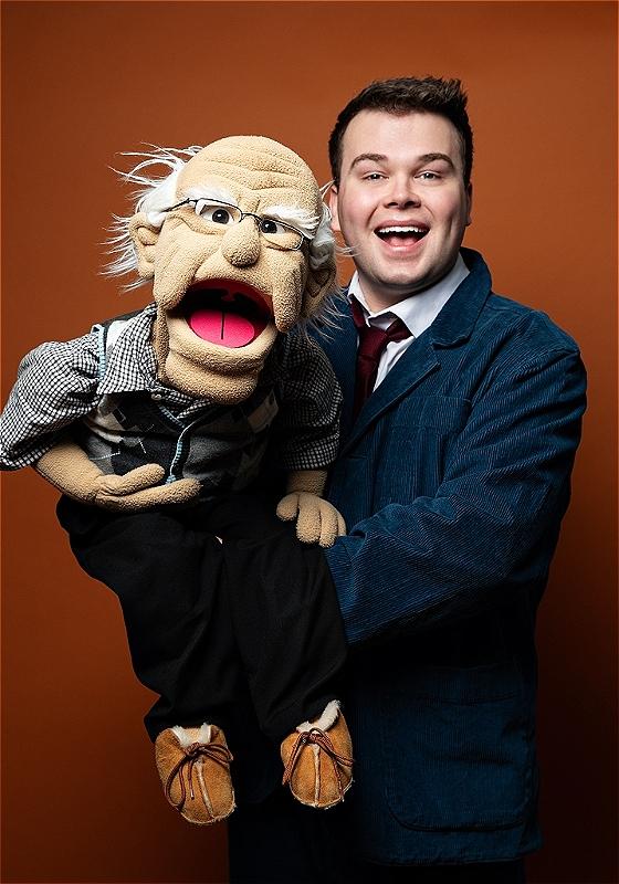Max Fulham stands wearing a shirt, tie, and blue suit jacket. He is standing against an orange background and holding a puppet of an old man with glasses.